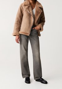 Veste en peau lainée marron avec col rabattable et fermeture éclair, associée à un jean droit gris clair et des mocassins noirs.