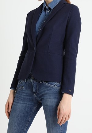 Nainen pukeutuneena istuvaan laivastonsiniseen bleiseriin, sinisen farkkupaidan ja keskiwash-väristen farkkujen kanssa valkoista taustaa vasten.