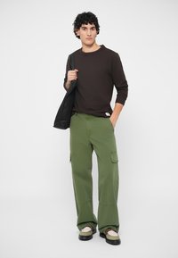Chemise marron à manches longues, pantalon cargo vert avec poches latérales, sac noir, chaussures beiges avec accents marron, design en tissu texturé.