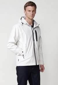 Lichte witte jack met een capuchon, voorzien van zwarte ritsen en accenten, een ritszak op de borst en verstelbare manchetten. Glad textuur.