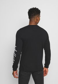 Svart långärmad T-shirt i bomull med rund hals och vit text längs vänster ärm. Slät textur, avslappnad design.