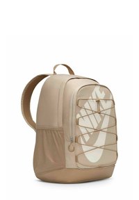 Mochila beige de Nike con un gran logo blanco, bolsillo lateral de malla y detalle de cordón elástico en la parte frontal. Tejido suave y correas acolchadas.