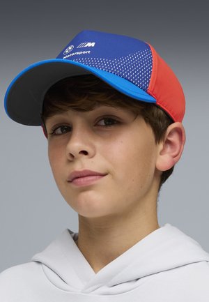 Giovane che indossa un cappellino BMW Motorsport blu e rosso e una felpa con cappuccio grigio chiaro su uno sfondo neutro.