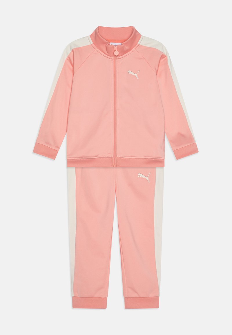 Roze trenirka s jaknom na zip i odgovarajućim hlačama. Ima bijele bočne pruge i logo na prsima i nogama. Izrađena je od mekog, glatkog materijala.