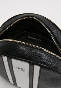 Borsa in pelle nera texturizzata con hardware argento, dotata di chiusura a zip e dettagli a righe bianche. L'interno mostra il marchio del designer.