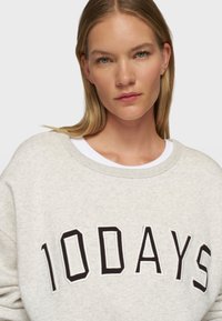 Sudadera gris claro con cuello redondo, que presenta un bold texto bordado en negro "10DAYS" en el pecho, hecha de una tela suave.