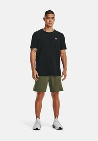Svart kortärmad idrottströja med en synlig logga, tillsammans med olivgröna shorts och vita sneakers, stående mot en vit bakgrund.