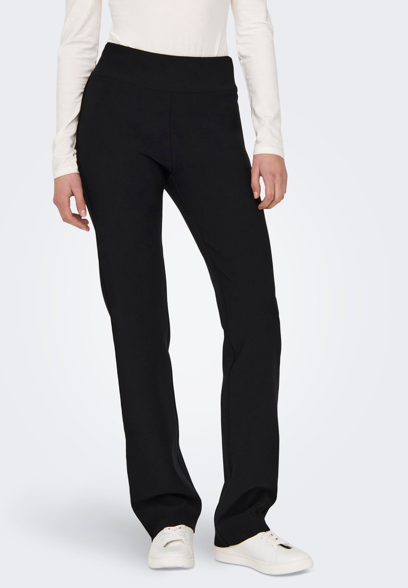 Sorte, rette benbukser laget av et elastisk stoff, med en høy, bred bærestropp for komfort. Kombinert med hvite joggesko.