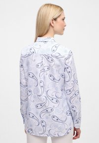 Camicia bianca con motivo paisley blu, maniche lunghe e polsini con bottone. Presenta un yoke posteriore dritto e un tessuto liscio e testurizzato.