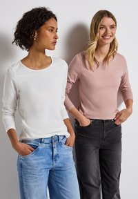 Twee vrouwen staan zij aan zij, gekleed in lange mouwen tops - de ene in het wit, de andere in lichtroze - gecombineerd met respectievelijk blauwe en zwarte jeans.