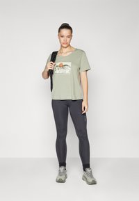 Ljusgrön t-shirt med kort ärm och en bergsgrafik samt en orange sol, i kombination med svarta leggings och grå vandringsskor.
