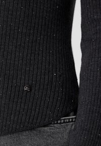 Pull en maille côtelée noire avec de petites paillettes argentées et une petite étiquette ronde en métal noir avec logo près de l'ourlet, porté avec un jean en denim gris.