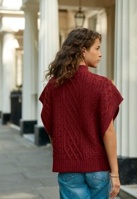 Poncho rouge tricoté avec un motif torsadé, ourlet côtelé, manches courtes, porté sur un jean en denim bleu. Dispose d'un col montant et d'une coupe ample.