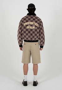 Personne portant une veste à carreaux marron et beige avec le texte « PROHIBITED BERLIN », un short beige, des chaussettes blanches, des chaussures noires et une casquette noire, debout.