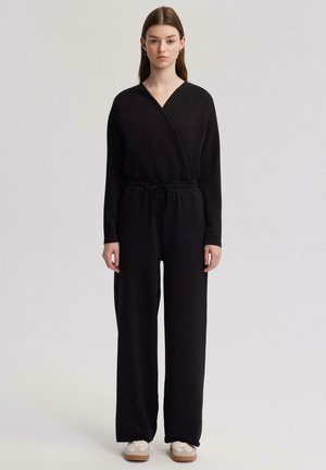 Sort jumpsuit lavet af blødt stof, med en indpaknings-overdel med V-udskæring, lange ærmer, elastisk talje og bred, lige ben-design.