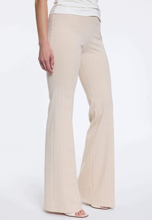 Beige pantalon met smalle strepen en wijde pijpen gedragen met witte sandalen met open teen en een wit topje, getoond van taille tot voeten.