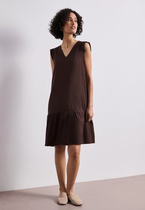 Mujer con cabello rizado, llevando un vestido sin mangas de color marrón oscuro y zapatos beige sin cordones, de pie frente a un fondo claro y liso.