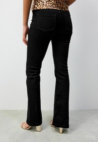Schwarze, ausgestellte Jeans aus Denim, mit einer taillierten Passform und auffälligen Gesäßtaschen. Getragen mit hellen High-Heels.