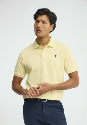 Hombre con una camisa tipo polo de color amarillo pálido y pantalones azul marino, de pie con las manos entrelazadas, mirando ligeramente hacia un lado contra un fondo simple.