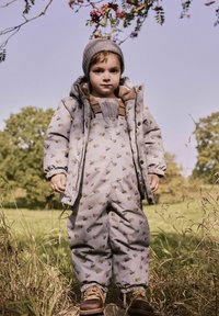 Młode dziecko ubrane w wzorzysty zimowy kurtkę, ogrodniczki, wełnianą czapkę i buty, stojące na zewnątrz w trawie na polu z drzewami i czystym niebem.
