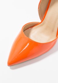 Chaussure à talon haut brillante, bout pointu orange, avec semelle intérieure beige rembourrée sur fond blanc.