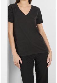 Svart v-ringad t-shirt tillverkad av mjukt, lätt material. Kort ärm och avslappnad passform. Buren med svarta byxor mot en neutral bakgrund.