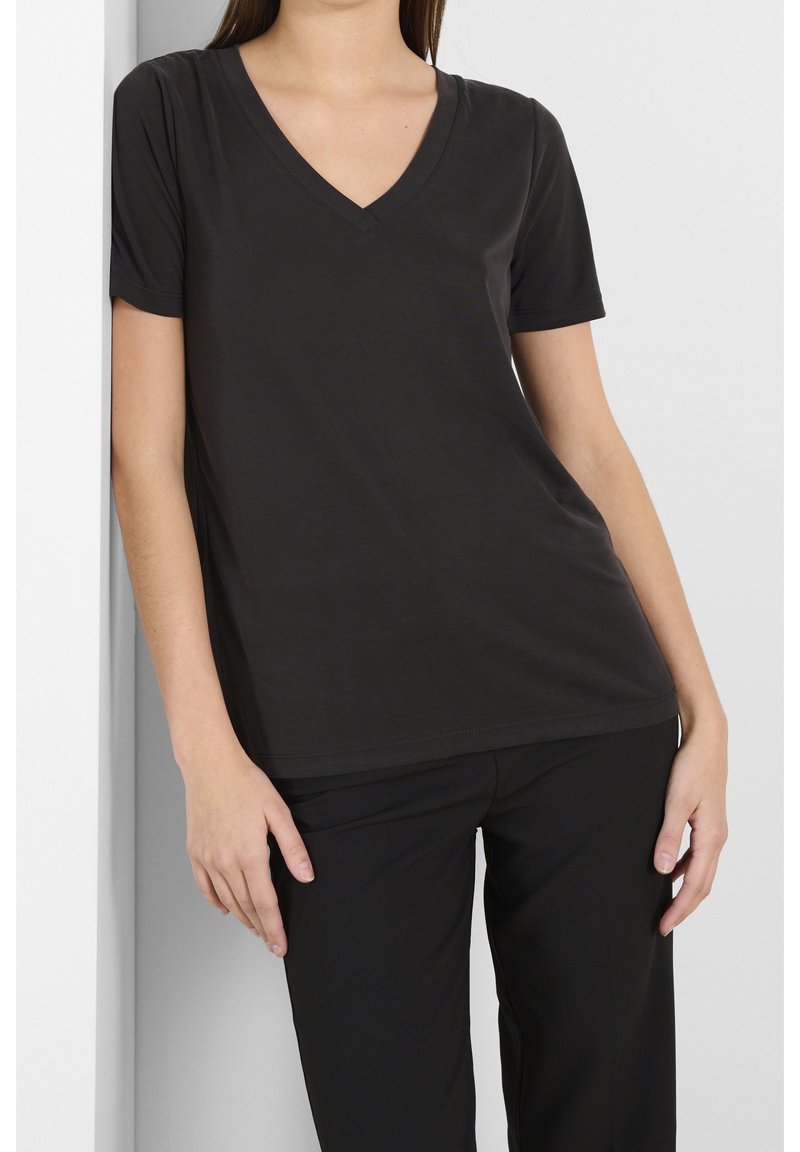 Svart v-ringad t-shirt tillverkad av mjukt, lätt material. Kort ärm och avslappnad passform. Buren med svarta byxor mot en neutral bakgrund.