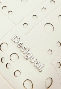 Accesorio de cuero blanco con recortes circulares, que presenta un logotipo plateado de "Desigual". La textura es suave con detalles de costura visibles.