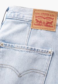 Vaaleansinisen denim-kankaan suorat farkut, joissa on nahkainen etiketti, eriväriset tikit ja metallinen nappi takataskussa.
