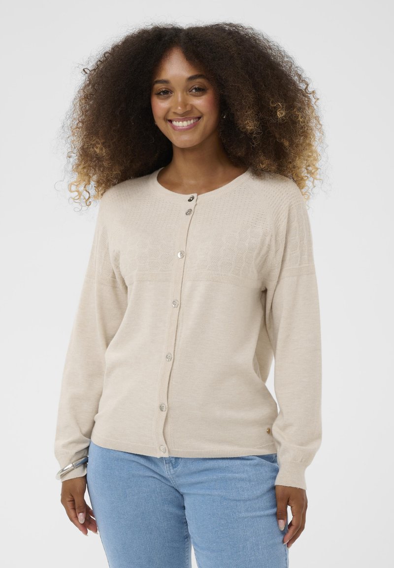 Nasmejana ženska oseba s kodrastimi lasmi, oblečena v bež kardigan s gumbi in svetleje modre kavbojke, na enotnem ozadju.