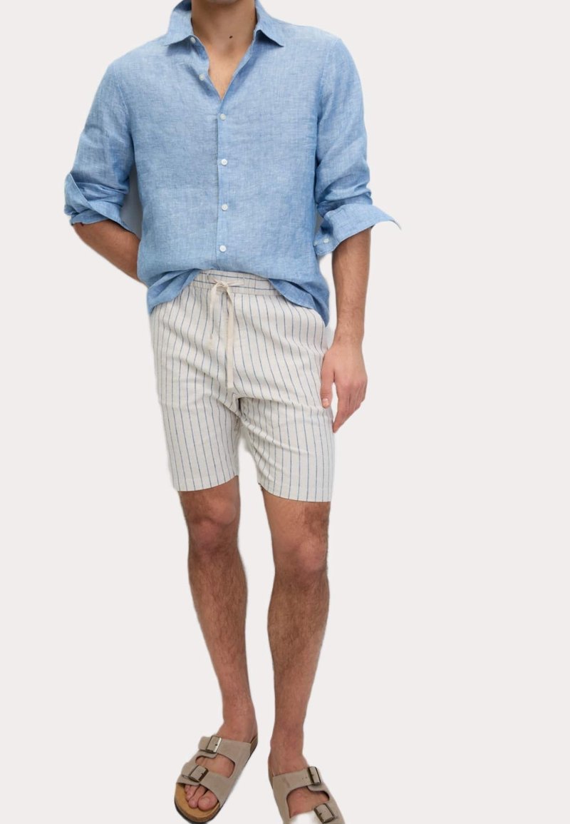 Homme portant une chemise bleu clair à manches longues, un short blanc rayé bleu avec cordon, et des sandales beiges à boucle, debout avec une main dans le dos.