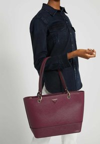 Sac fourre-tout synthétique bourgogne avec surface texturée, poignées doubles et quincaillerie en argent. Présente un logo triangulaire à l'avant et un design structuré.