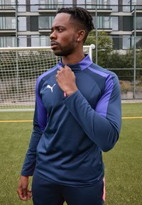 Karinė sportinė striukė su purpuriniais akcentais, aukšta apykakle ir užtrauktuku; ant krūtinės yra baltas logotipas, tekstūruotas audinys ir prigludęs dizainas.
