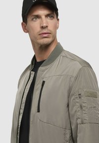 Khaki bomberjack met een rits, geribbeld kraag en gestructureerde schouderpatches. Onder de jas zit een zwart T-shirt en een zwarte pet.