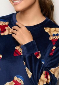 Maglione in morbido tessuto a maniche lunghe di colore blu navy con un motivo ripetuto di orsetti in peluche con cappelli e sciarpe nei toni del rosso e del blu. Texture soffice.