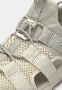 Ανοιχτό μπεζ σανδάλι με επάνω μέρος από δίχτυ, ελαστικό σύστημα κορδονιών και σχεδιασμό λουριού με υφή για αναπνοή και άνεση.