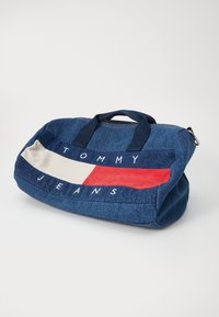 Sac de sport en denim avec des poignées bleu marine, présentant un design en blocs de couleurs horizontal de rouge, blanc et bleu. Logo "TOMMY JEANS" affiché de manière proéminente.
