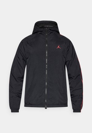 Veste de sport noire avec capuche, présentant des accents rouges sur les manches et un logo rouge sur la poitrine, fabriquée en matériau léger.