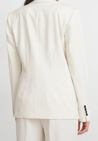 Wit, op maat gemaakte blazer met een gladde textuur, gestructureerd ontwerp, enkele achtersplit en zwarte knopen op de manchetten.