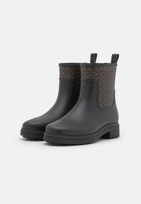 Une paire de bottines noires à la cheville avec un panneau en tissu monogrammé marron et des languettes de tirage sur fond blanc uni.
