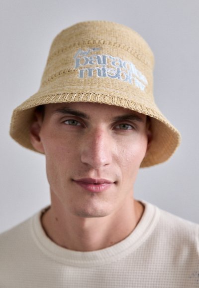 Chapeau seau en paille tissée de couleur brun clair, orné de texte brodé bleu clair. Le chapeau possède une large visière et une surface texturée, porté par une personne.