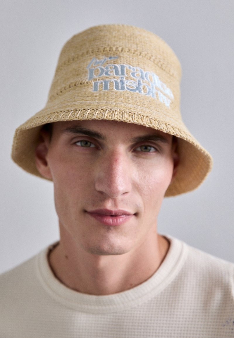Tan woven straw bucket hat featuring light blue embroidered text. Hat has a wide brim and textured surface, worn by a person.