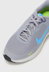 Chaussure de sport gris clair avec une tige en mesh respirant, accents bleus et verts fluo, bout arrondi et système de laçage traditionnel.