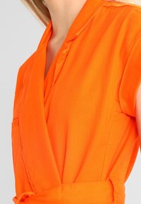 Lys orange omslagkjole med korte ærmer, reverskrave og bælte i taljen. Har en lomme på venstre side og en glat stofstruktur.