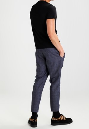 Mężczyzna w czarnym t-shircie, niebieskich kraciastych spodniach 3/4, czarnych skarpetkach i czarnych butach z detalami w panterkę, stoi z rękami w kieszeniach.
