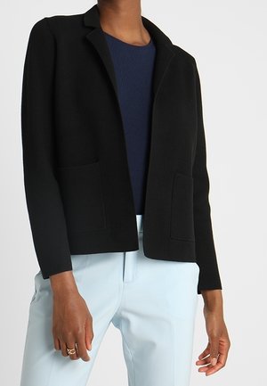 Donna che indossa un blazer nero sopra un top blu navy e pantaloni azzurri, in piedi con le braccia rilassate lungo i fianchi.