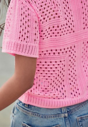 Top rosa all'uncinetto con un design a trama larga e aperta, maniche corte e orlo dritto, abbinato a pantaloni shorts di denim sullo sfondo.