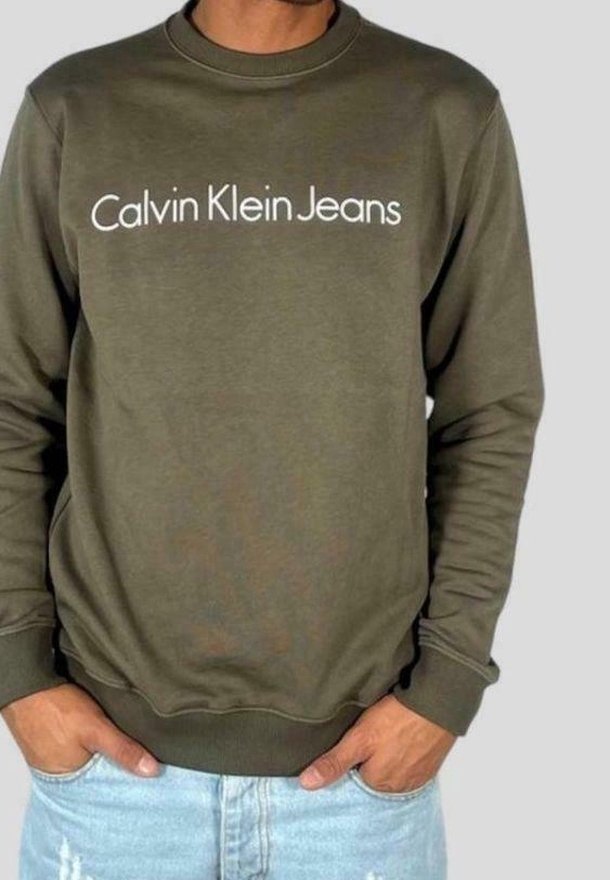Homme portant un sweat-shirt Calvin Klein Jeans vert olive, mains dans les poches, associé à un jean bleu clair déchiré.