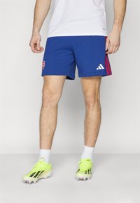 Mėlyni sportiniai šortai su raudonomis šoninėmis juostelėmis ir Arsenal logotipu, derinami su ryškiai geltonais futbolo batais ir baltais kojinės.