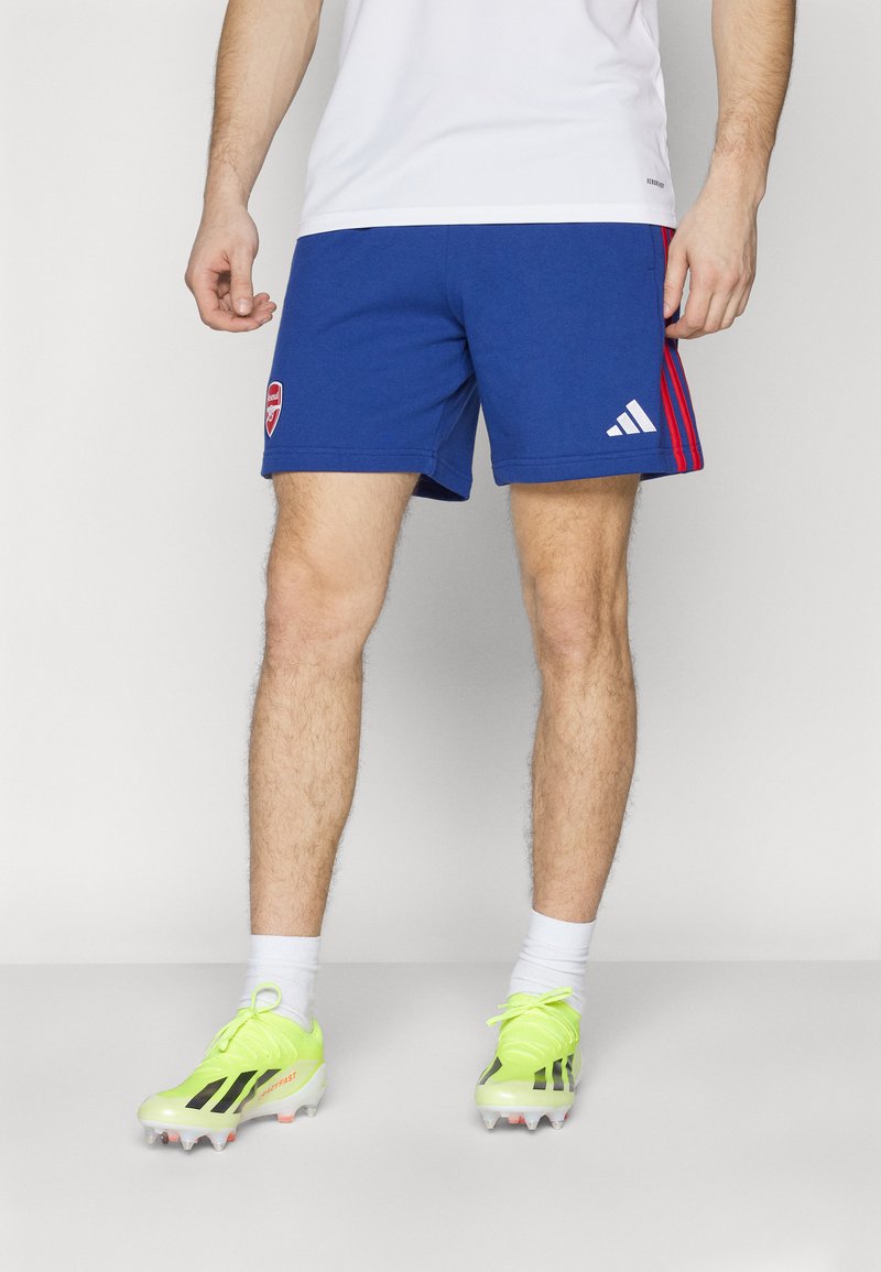 Mėlyni sportiniai šortai su raudonomis šoninėmis juostelėmis ir Arsenal logotipu, derinami su ryškiai geltonais futbolo batais ir baltais kojinės.